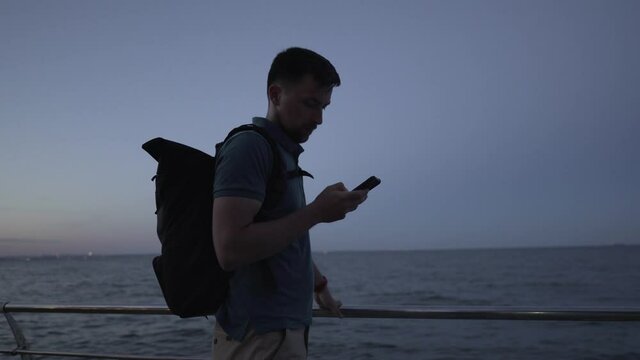 Man takes pictures in evening twilight of natural environment of sea using modern smartphone camera, guy traveler with backpack holding mobile phone, making photo sea water through an application.