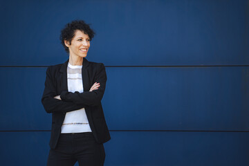 Smiling mature businesswoman looking at copy space while standing arms crossed against blue wall