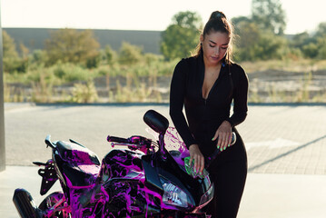 Fototapeta na wymiar Beautiful girl in tight-fitting seductive suit washes black sport motorcycle and wipes it from magenta foam at sunrise.