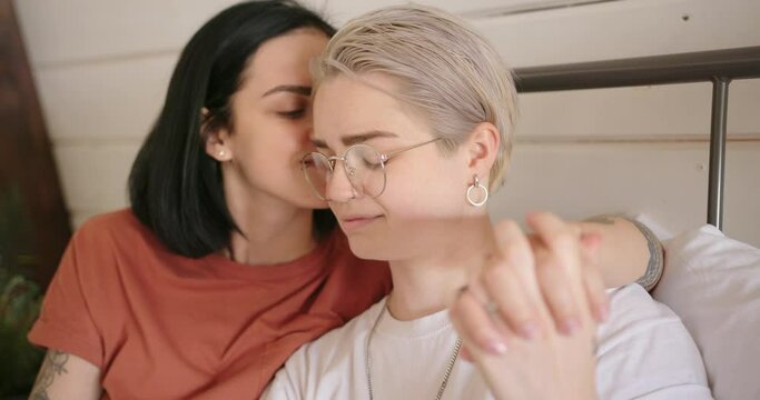 Smiling brunette and blonde with tattoos join hands and kiss lying on bed at home with white wall slow motion close view
