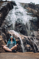 Young white girl sitting under Diyaluma waterfall, Sri Lanka. Happy woman in meditation pose on vacation. Wellness lifestyle, healthy yoga retreat in summer. Peaceful holiday in beautiful exotic place