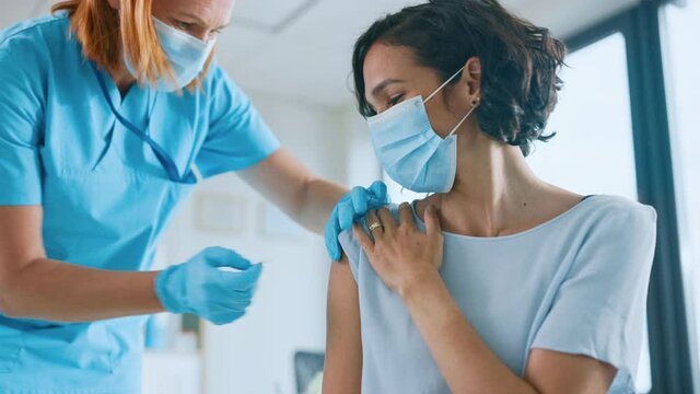 Medical Nurse in Safety Gloves and Protective Mask is Making a Vaccine Injection to a Female Patient in a Health Clinic. Doctor Uses Hypodermic Needle and a Syringe to Put a Shot of Drug as Treatment.