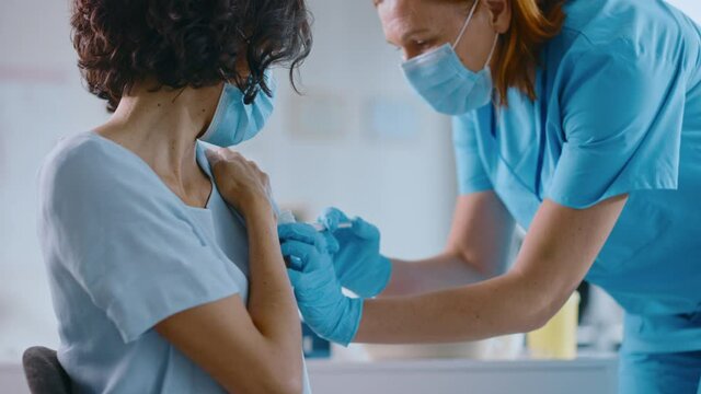 Medical Nurse in Safety Gloves and Protective Mask is Making a Vaccine Injection to a Female Patient in a Health Clinic. Doctor Uses Hypodermic Needle and a Syringe to Put a Shot of Drug as Treatment.