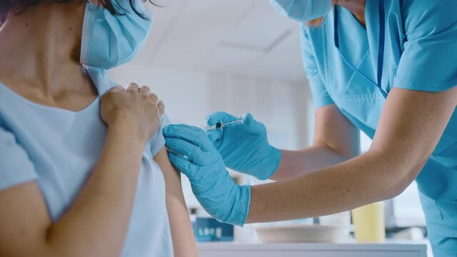 Medical Nurse in Safety Gloves and Protective Mask is Making a Vaccine Injection to a Female Patient in a Health Clinic. Doctor Uses Hypodermic Needle and a Syringe to Put a Shot of Drug as Treatment.