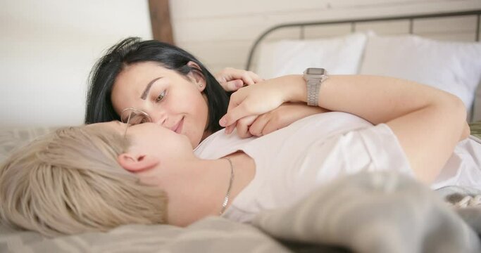 Lesbians with tattoos of black colour lie on bed and kiss looking into eyes against white wall at home slow motion close view