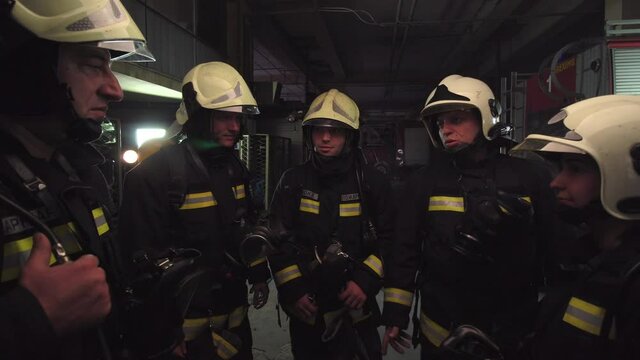 Group of five firefighters wearing full equipment in smokey area. Relaxed chatting after work well done. Fire trucks in the background. Name tag translation is fireman and model's last name.