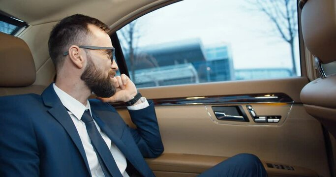 Handsome Caucasian happy man in glasses sitting on backseat of expensive car and talking on cellphone. Successful cheerful businessman speaking on moile phone. Telephone call. Business conversation.