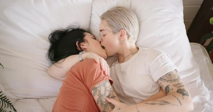 Romantic lesbian couple with tattoos hugs and kisses lying on bed with large pillows between pot plants slow motion close view