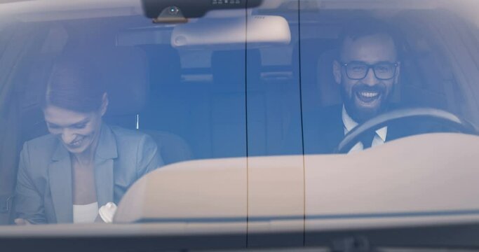 Happy cheerful Caucasian wife and husband driving early in morning to work and laughing. Man driving and woman using mobile, drinking coffee. Couple of business colleagues in automobile. Sipping drink
