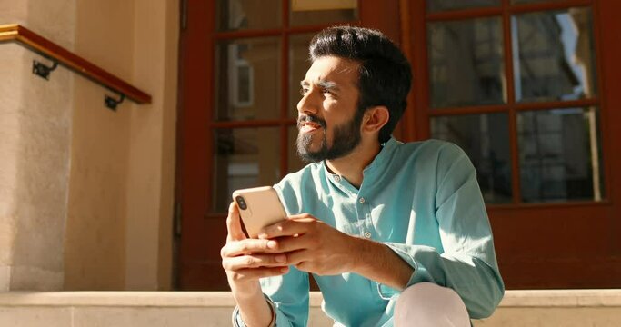 Hindu young handsome man in traditional outfit sitting on steps and texting message on smartphone. Sms sending. Guy tapping and scrolling on mobile phone. Male messaging outdoor. India concept.