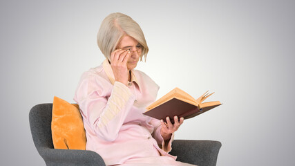 Retired woman reading a book sitting on a chair on gradient back