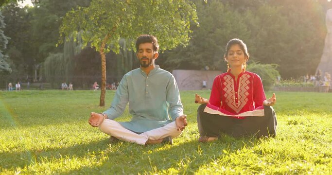 Hindu couple of man and woman sitting in lotus pose on green grass and meditating. Male and female yogis in traditional clothes in park on sunny day having meditation. Yoga outdoor in summer in India.
