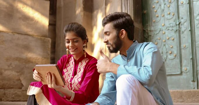 Happy cheerful young Hindu man and woman sitting outdoor at steps and using tablet device while videochatting. Videochat on gadget computer. Male and female talking via web cam.