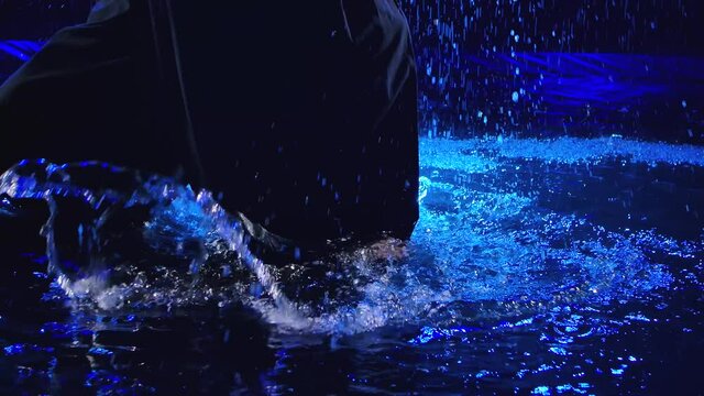 Legs of a kendo fighter in a black kimono practicing movement on the water surface under the rain and splashes of water. Close up. Slow motion.