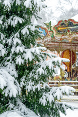 Obraz premium A fabulous view of a colorful carousel through a snow-covered spruce branch during a snowfall in the park.