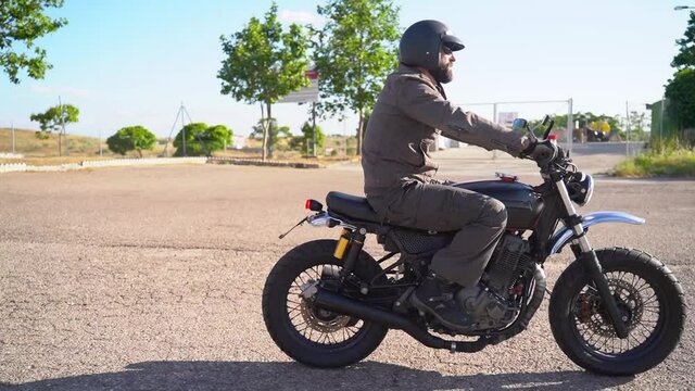 Tracking side shot for an adult man with beard and helmet riding modern black moto bike on asphalt road during the day