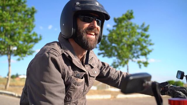 Approaching a dressed beard biker with helmet and sun glasses siting on a motorcycle and smiling. blue sky in background.