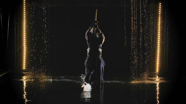 A Japanese kendo fighter wearing armor and traditional kimono practices a martial art with a bamboo bokken against a black background in the rain. Slow motion.
