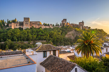 Naklejka premium Granada, Spain. October 17th, 2020. View at sunset from below of the buildings and towers of the Alhambra, with a palm tree and some house roofs in the foreground.