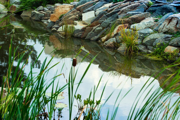 Beautifully decorated with stones and plants artificial body of water with a reflection of the sky on the water.