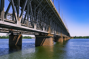 Powerful Amur bridge over Dnieper river in Dnipro (Ukraine). Steel structure with concrete support beams in the water. Beautiful architectural engineering background with copy space and perspective