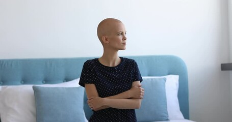 Female cancer patient sit on bed alone with arms-crossed looking out the window. Bald young woman feels afraid of upcoming operation. Struggle for life, oncology disease clinic cancer patient concept