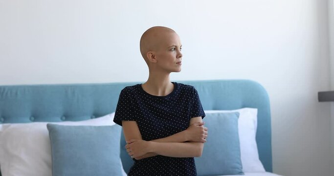Female cancer patient sit on bed alone with arms-crossed looking out the window. Bald young woman feels afraid of upcoming operation. Struggle for life, oncology disease clinic cancer patient concept