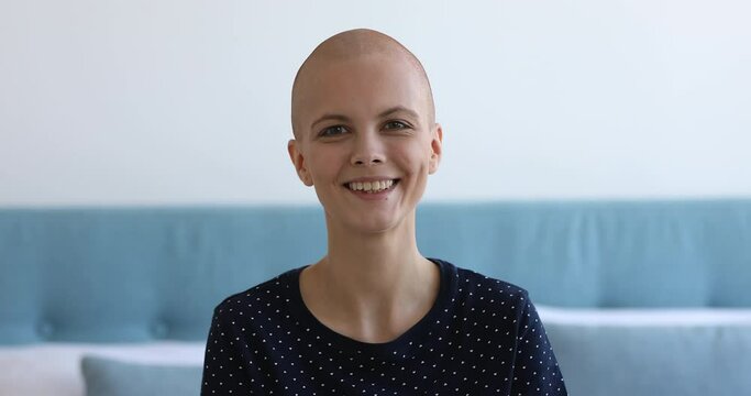 Head shot portrait pretty young bald woman sit on bed in bedroom alone smiling looks away feels hopefulness and confidence about the healthy future and success of innovative cancer treatments concept