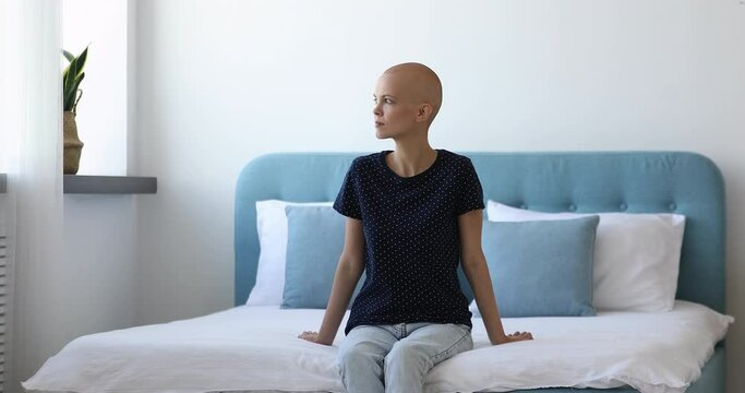 Pensive cancer patient young bald woman sitting in bedroom on bed looking out the window smiles and daydreams, hopes sincere believes for recovery. Oncology treatment center 30s female patient concept