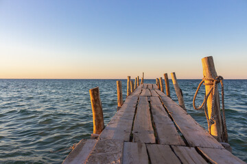 Wooden pier at the sunset. Evening sky over sea with footbridge. Calm evening landscape. Empty pier at seaside with copy scape. Pier in warm sunset light. Horizon over sea. Marine travel. 
