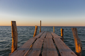 Fototapeta na wymiar Wooden pier at the sunset. Evening sky over sea with footbridge. Calm evening landscape. Empty pier at seaside with copy scape. Pier in warm sunset light. Horizon over sea. Marine travel. 