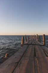 Fototapeta premium Wooden pier at the sunset. Evening sky over sea with footbridge. Calm evening landscape. Empty pier at seaside with copy scape. Pier in warm sunset light. Horizon over sea. Marine travel. 
