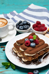 portion of Classic tiramisu dessert with raspberries and blueberries and cup of espresso coffee on blue wooden background