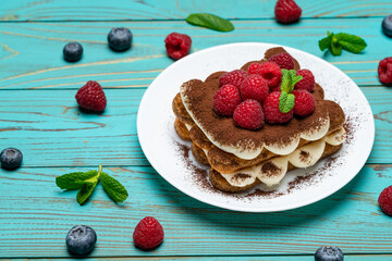 portion of Classic tiramisu dessert with raspberries and blueberries on blue wooden background