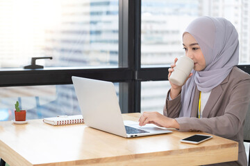 Confident Asia Muslim (Islam) businesswomen executive working in the modern office drinking a cup of coffee and looking on a laptop and sit beside windows. Work concept of diversity of culture..