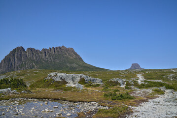 Wide angle view on the Overland Track landscape. Hiking trail, green grass, and shallow pond shallow against a background of Cradle Mountain and another steep and rocky mountain. Tasmania, Australia
