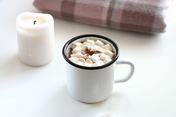 Hot cocoa with marshmallows in a white ceramic mug on a white table. Nearby lies a warm blanket and a candle.