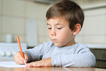 Boy in the kitchen at the table draws with a pencil on a sheet of paper