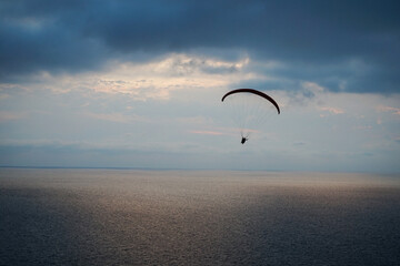 a paraglider flies over the sea at sunset against the background of the light part of the sky between the dark sea and dark clouds