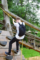 Tennage boy climbs on wooden climbing wall with ropes outdoors in adventure rope park, back view. Outdoor activities for healthy lifestyle.

