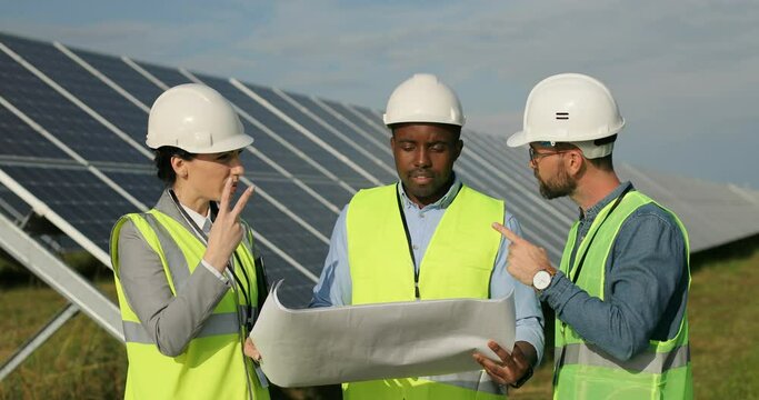 Three technical engineers review plan on paper and discuss it by gesturing with hands. Solar plantation. Multinational team.