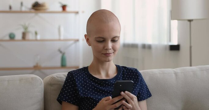 Bald woman sit on couch with smartphone enjoy fulfilling life, smiles texting message use social media network chat resting alone at home. Oncology disease cancer patient and modern tech usage concept