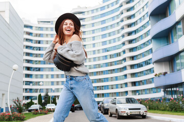 Outdoor image of joyful girl in black wool hat and grey fall sweater jumping and enjoying walk in modern urban city.