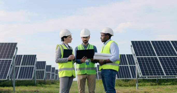 Three engineers in unifom: one woman and two men standing among solar panels and discussing layout of facilities.