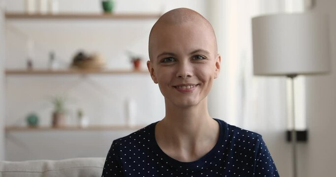 Head shot young bald woman sitting on couch smiling look at camera. Cancer patient and victory over oncology disease, hopeful ill hairless caucasian girl portrait. Rehabilitation and remission concept