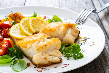 Fish dish - fried cod fillet with fried vegetables and lemon served on wooden table