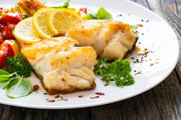 Fish dish - fried cod fillet with fried vegetables and lemon served on wooden table