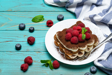 portion of Classic tiramisu dessert with raspberries and blueberries on blue wooden background
