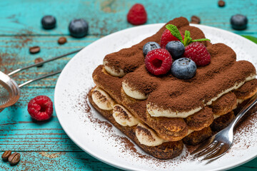 portion of Classic tiramisu dessert with raspberries and blueberries on blue wooden background