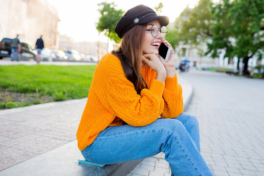 Outdoor lifestyle image of romantic   dreamy woman sitting on sidewalk and tolking by mobyle phone . Wearing stylish jeans and orange sweater.